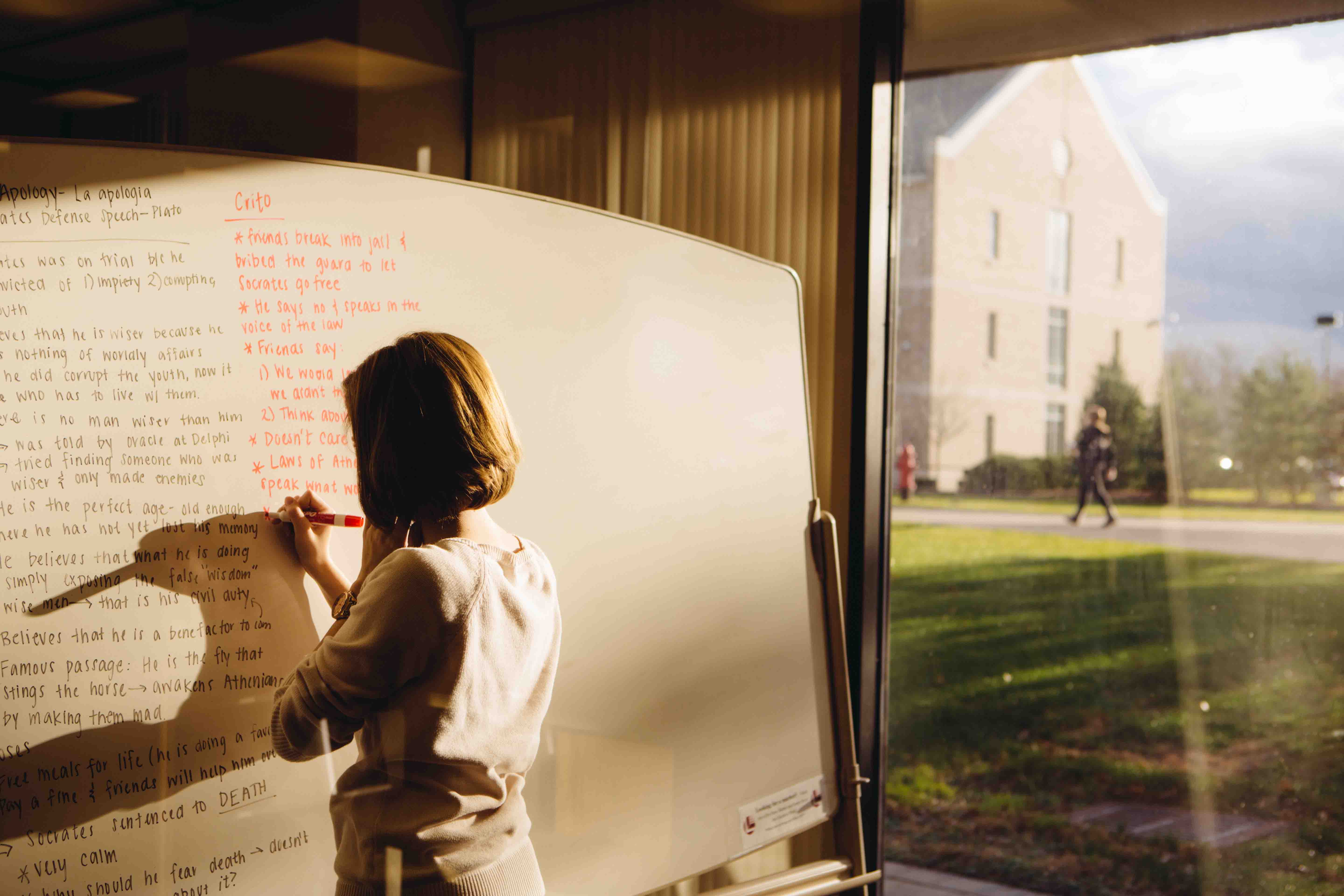 College student writing on a white board with campus in background.